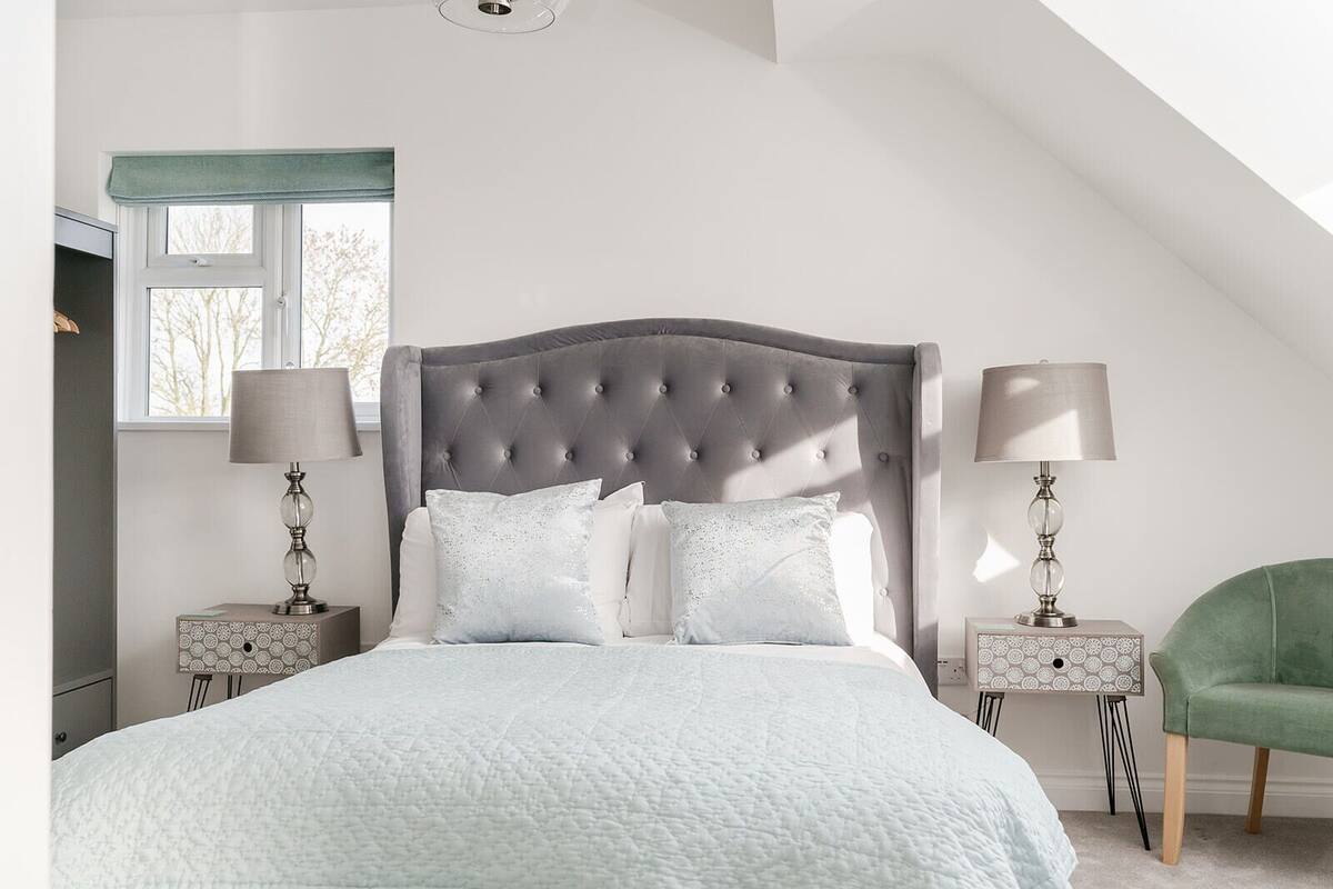 A bedroom with a large bed, two bedside tables with lamps, a green chair, and a window with a view of trees.