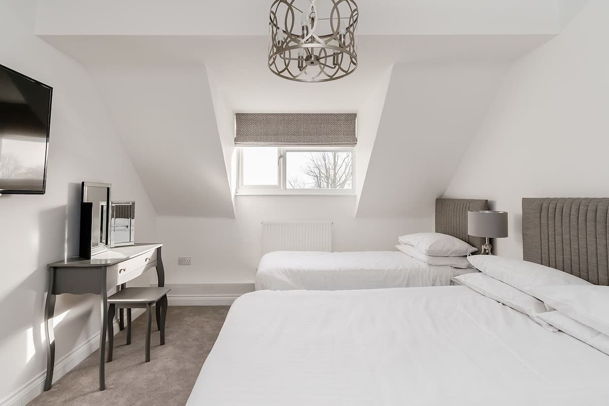 A modern, minimalist bedroom with a sloped ceiling, a large window, a desk with a chair, two beds, and a chandelier.