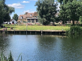 A calm lake with a dock, surrounded by greenery and a large house in the background.