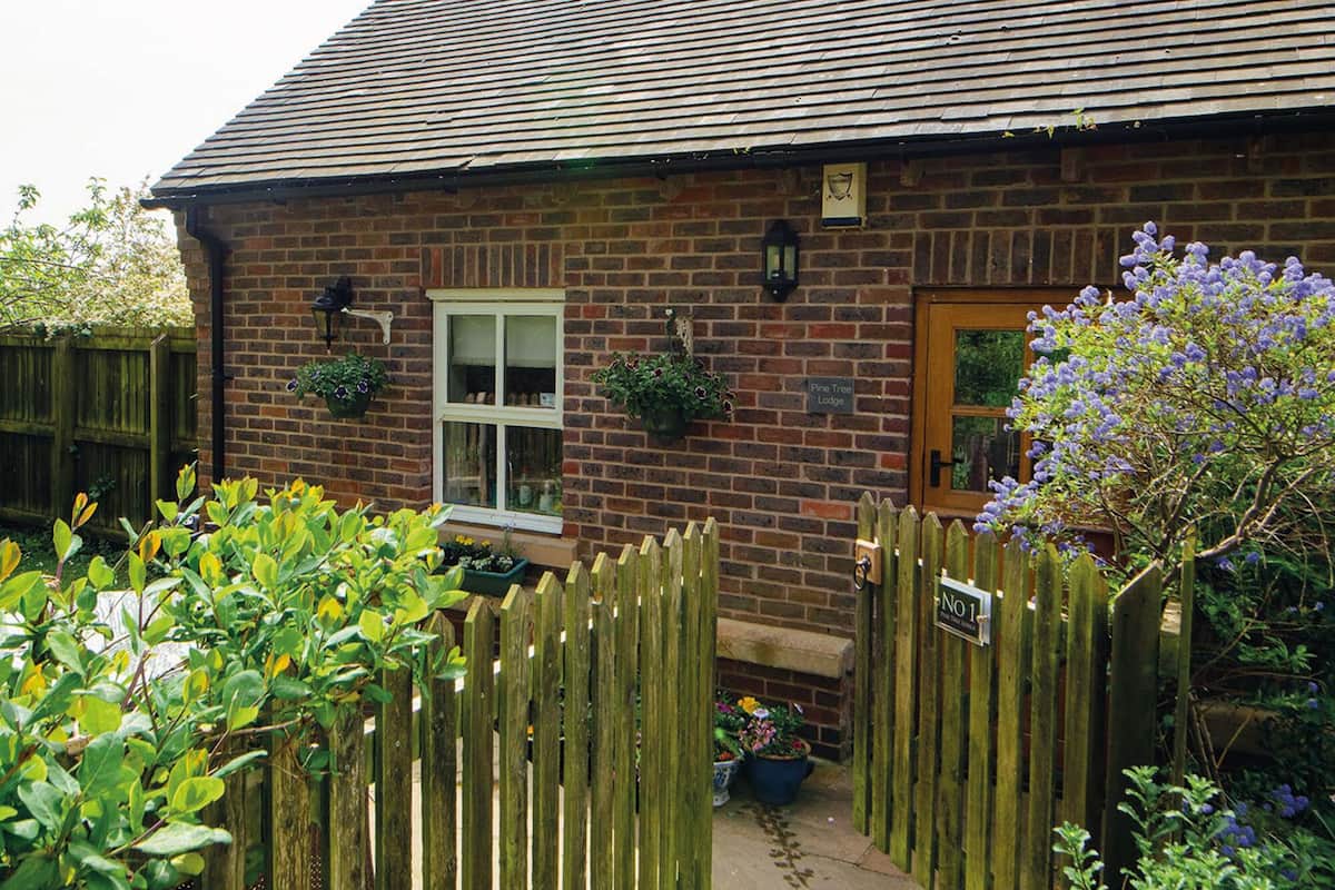 A brick cottage with a wooden fence, a window with hanging flower pots, and a sign reading "Pine Tree Lodge."