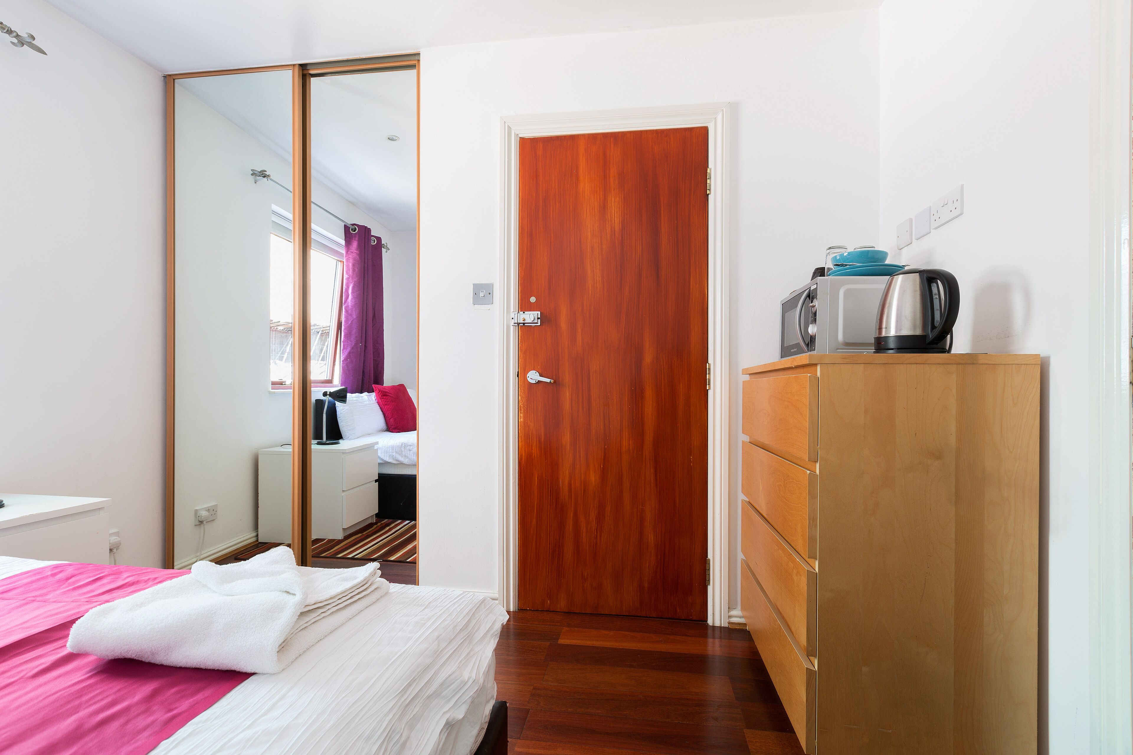 A hotel room with a wooden wardrobe, a bed with a pink and white bedspread, a wooden floor, and a door leading to another room.