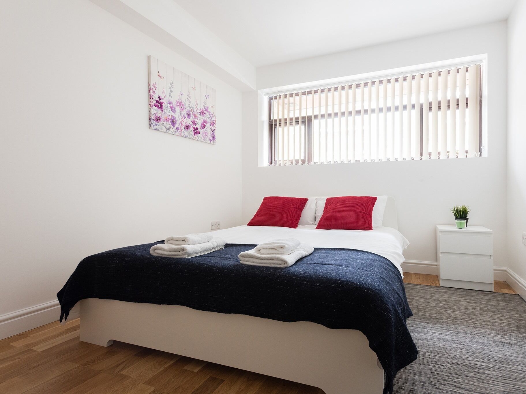 Un dormitorio ordenado con cama, almohadas, una mesita de noche y una ventana con persianas.