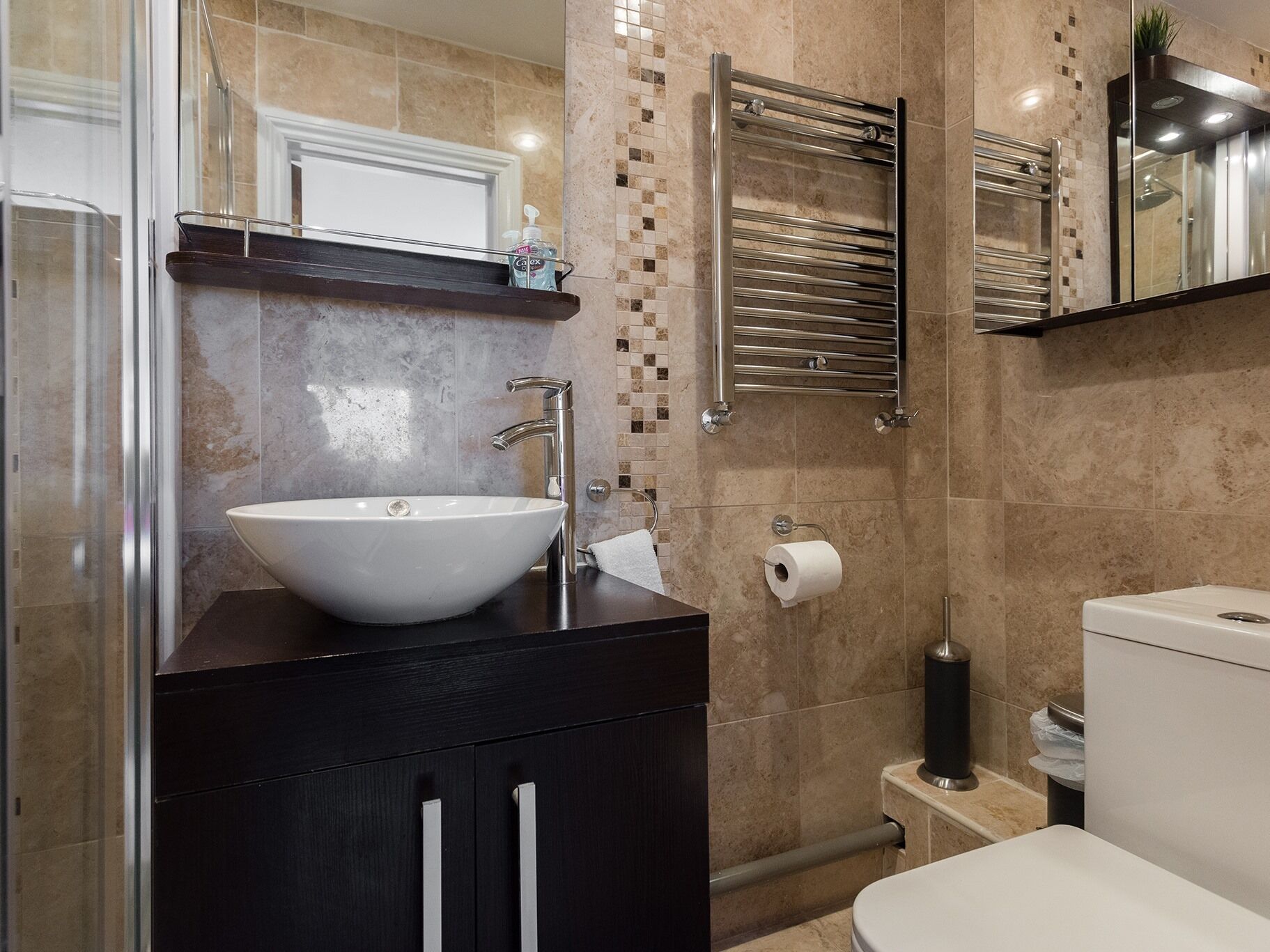 A modern bathroom with a white sink, dark cabinetry, a glass shower enclosure, a towel rack, and a toilet.