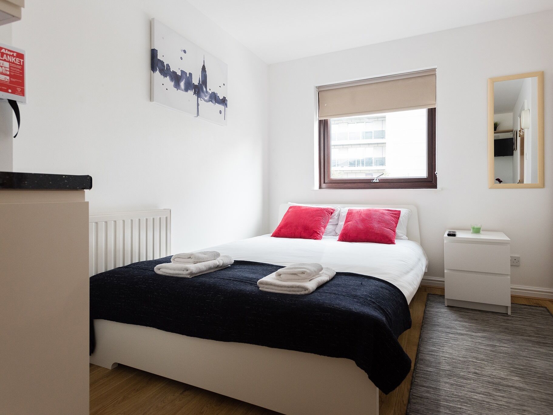 Habitación de hotel ordenada con cama, mesita de noche, ventana con vistas, radiador y una obra de arte colgada en la pared.