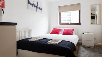Habitación de hotel ordenada con cama, mesita de noche, ventana con vistas, radiador y una obra de arte colgada en la pared.