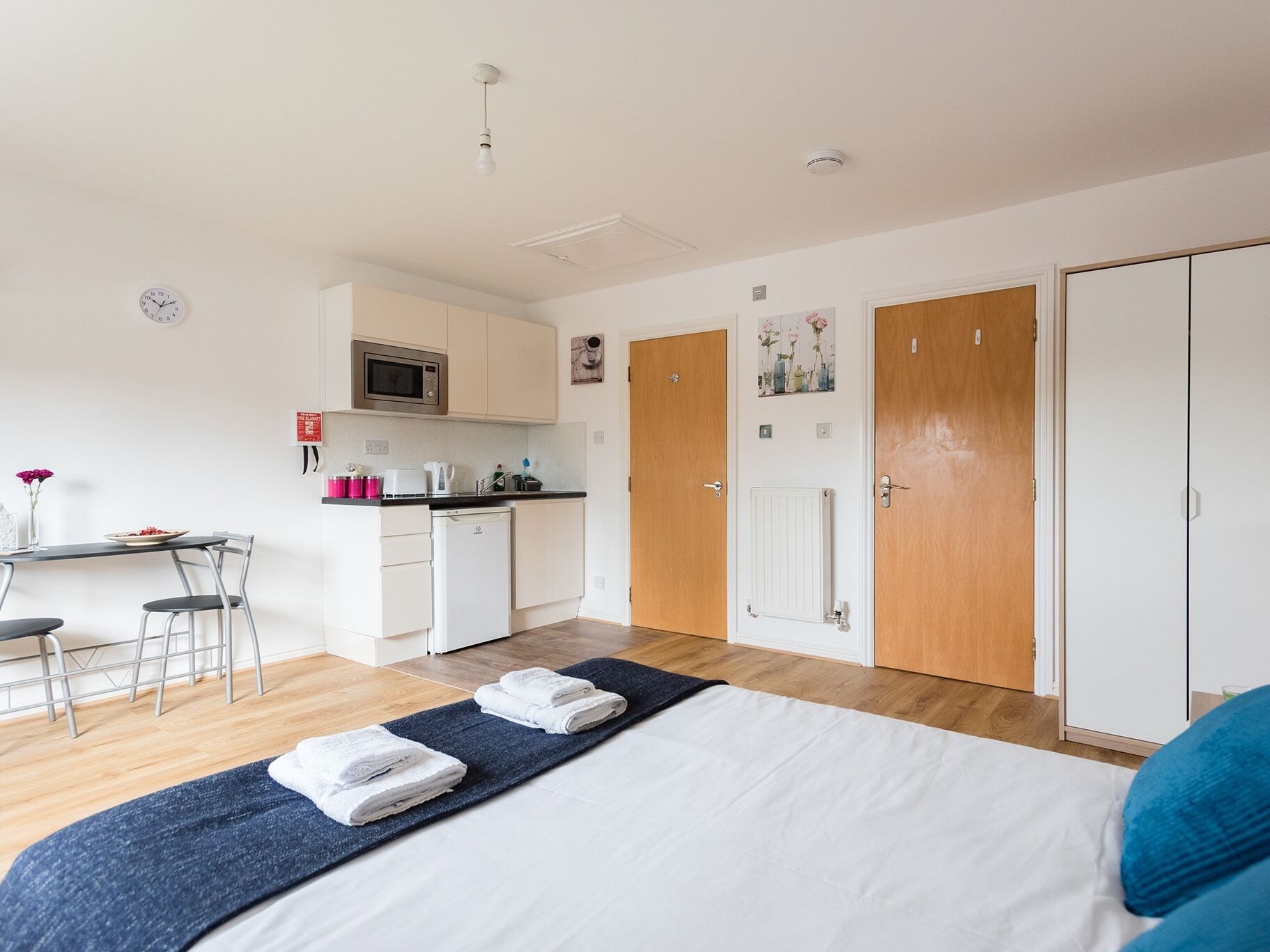 A modern hotel room with a bed, a desk, a microwave, a small kitchenette, and a wardrobe.