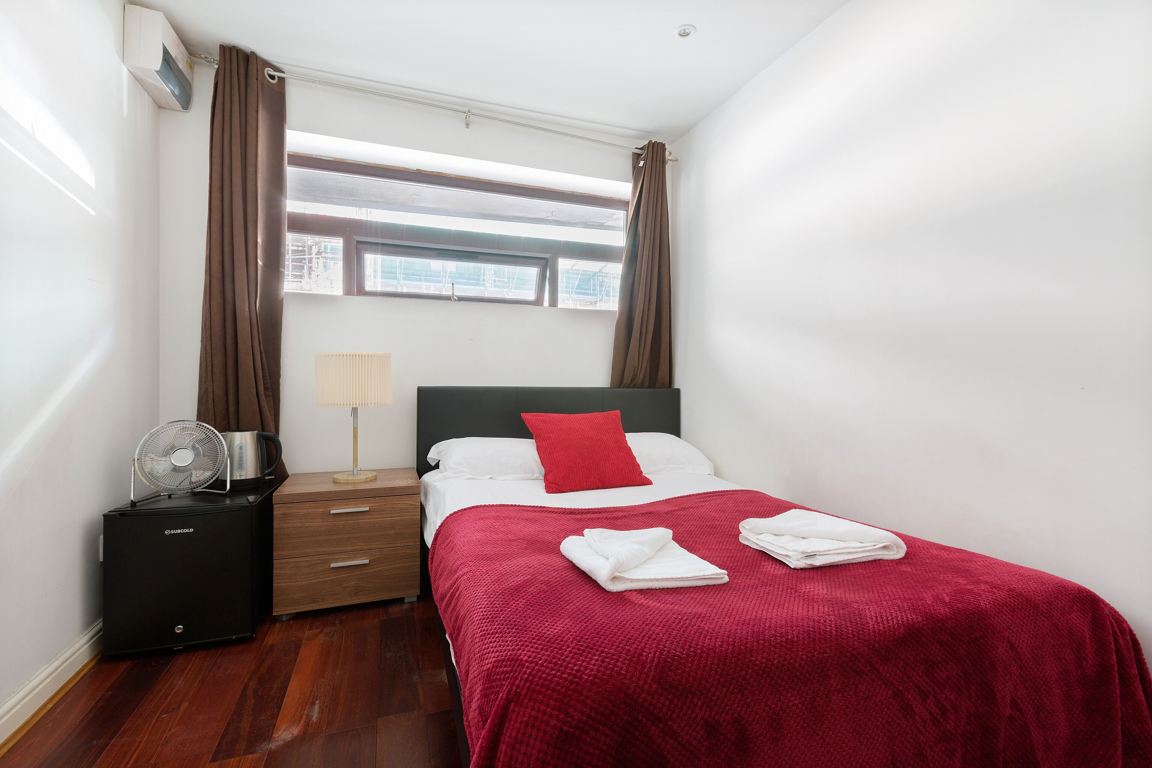 A hotel room with a bed, a bedside table, a fan, and a window with curtains.