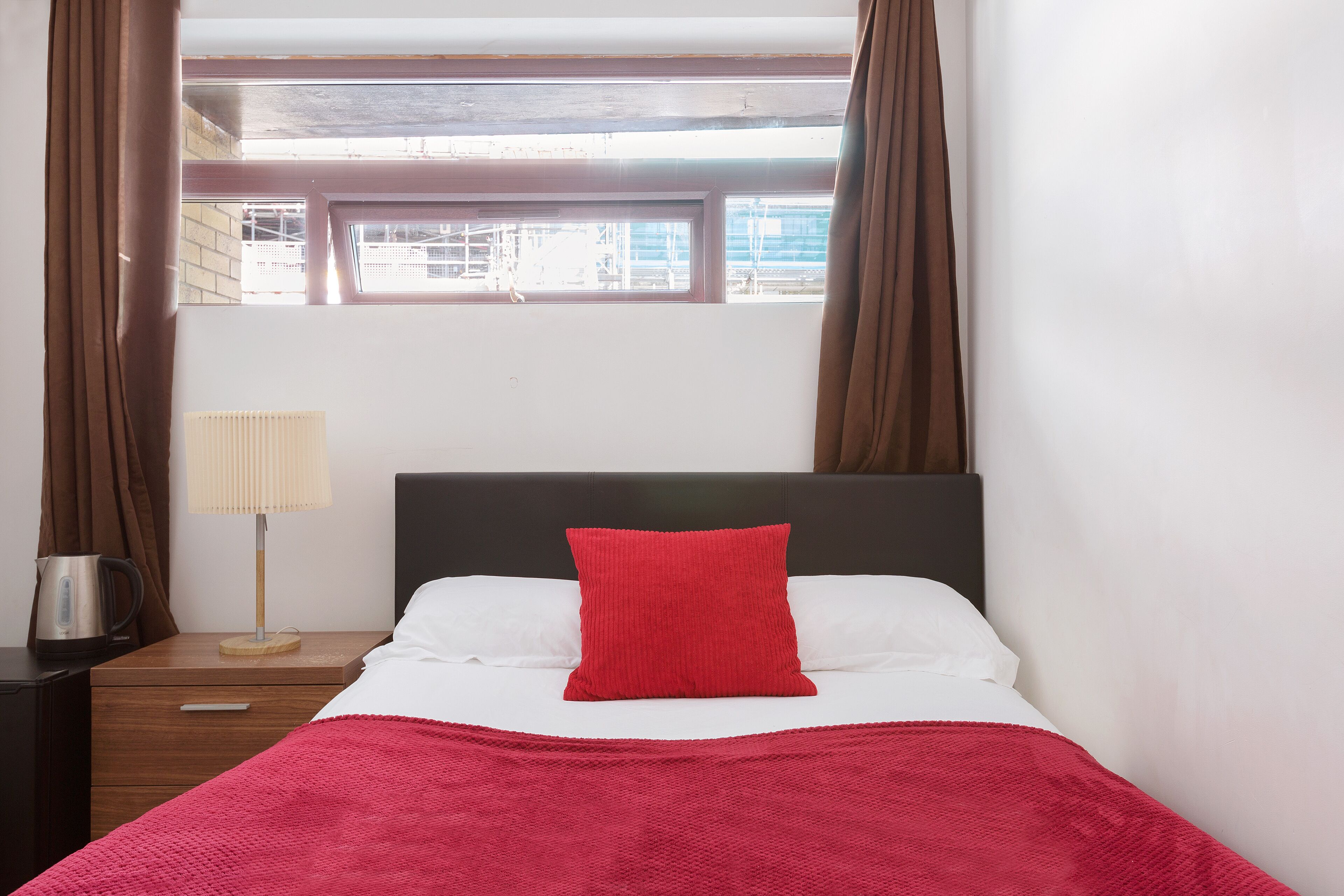 A bed with a red pillow, white sheets, and a red blanket. There is a nightstand with a lamp and a kettle. The room has a window with curtains.