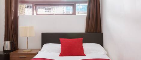 A bed with a red pillow, white sheets, and a red blanket. There is a nightstand with a lamp and a kettle. The room has a window with curtains.