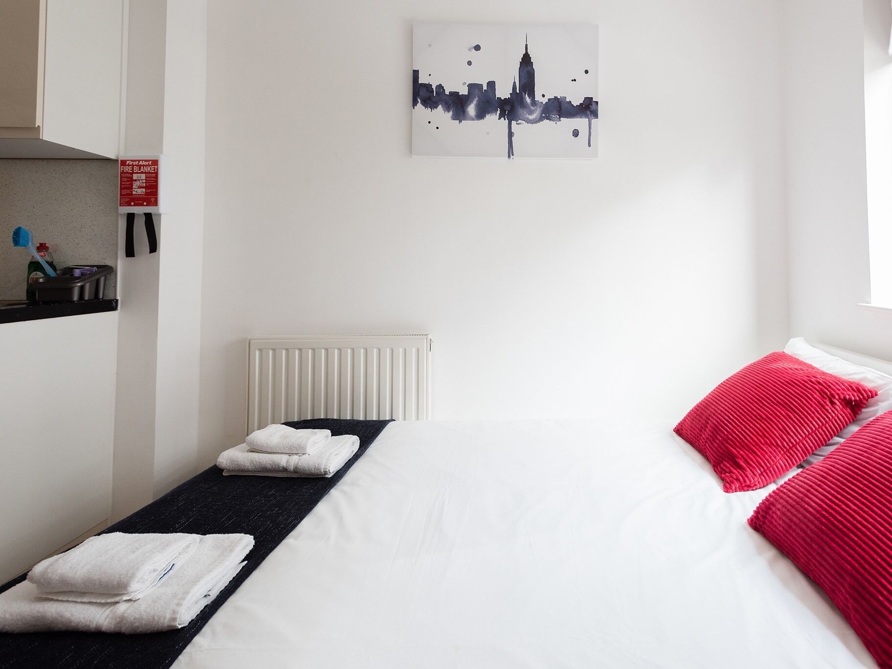 Habitació d'hotel amb un llit, un radiador, una manta ignífuga d'auxili i una obra d'art amb el paisatge urbà a la paret.