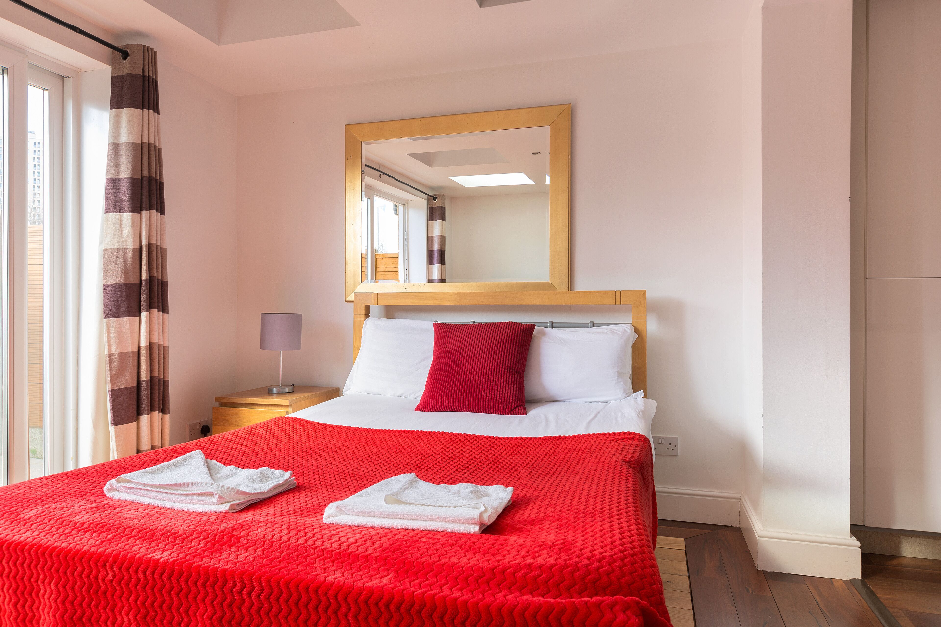 Habitación de hotel con cama, manta roja, almohada blanca, almohada roja, cabecera de madera, espejo, mesita de noche, lámpara y ventana con cortinas.
