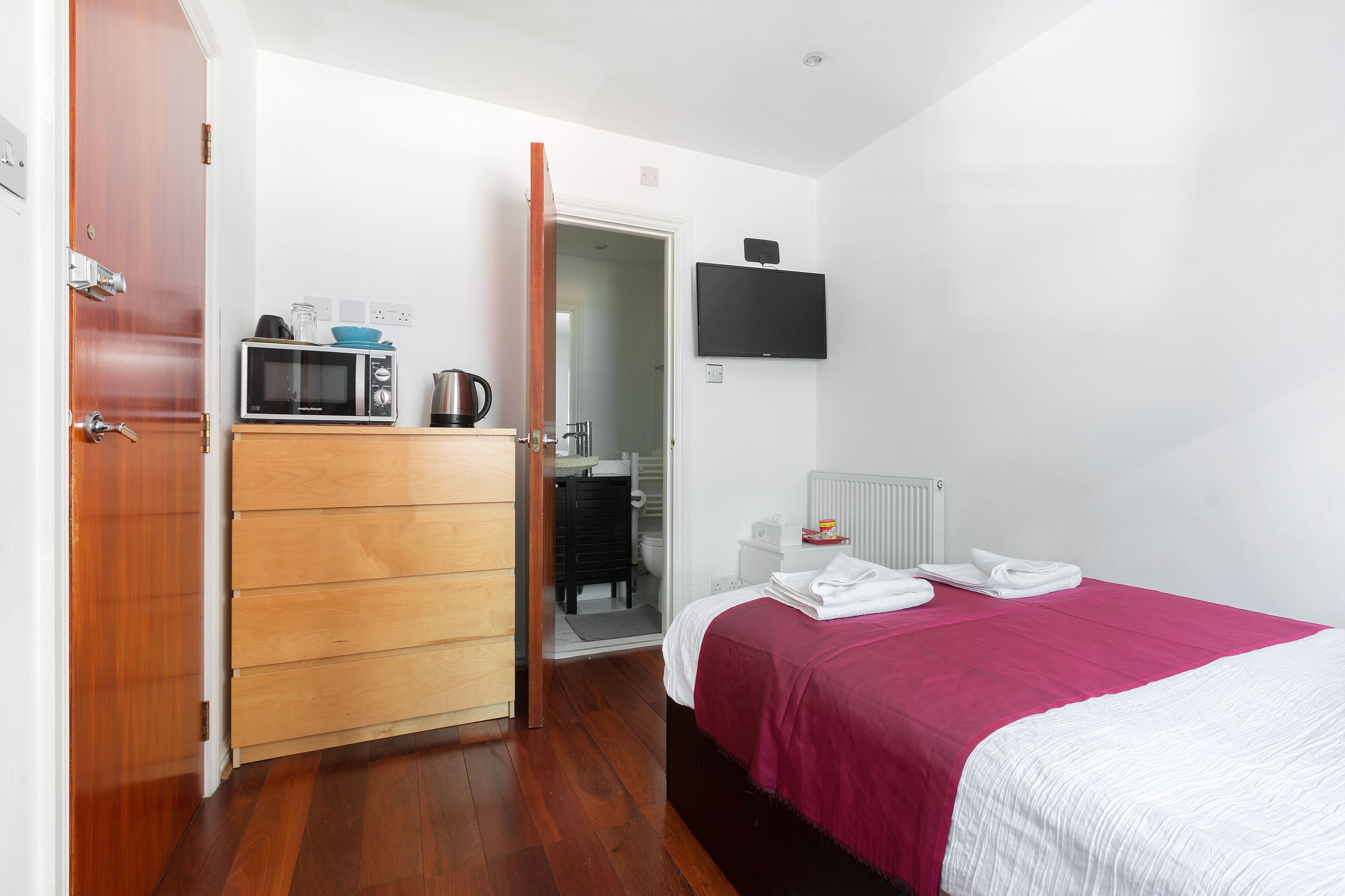 Une chambre d’hôtel avec une armoire en bois, un lit recouvert d’une couvre-lit rose et blanc, un four à micro-ondes posé sur la commode, un téléviseur accroché au mur et une salle de bain visible à travers une porte ouverte.