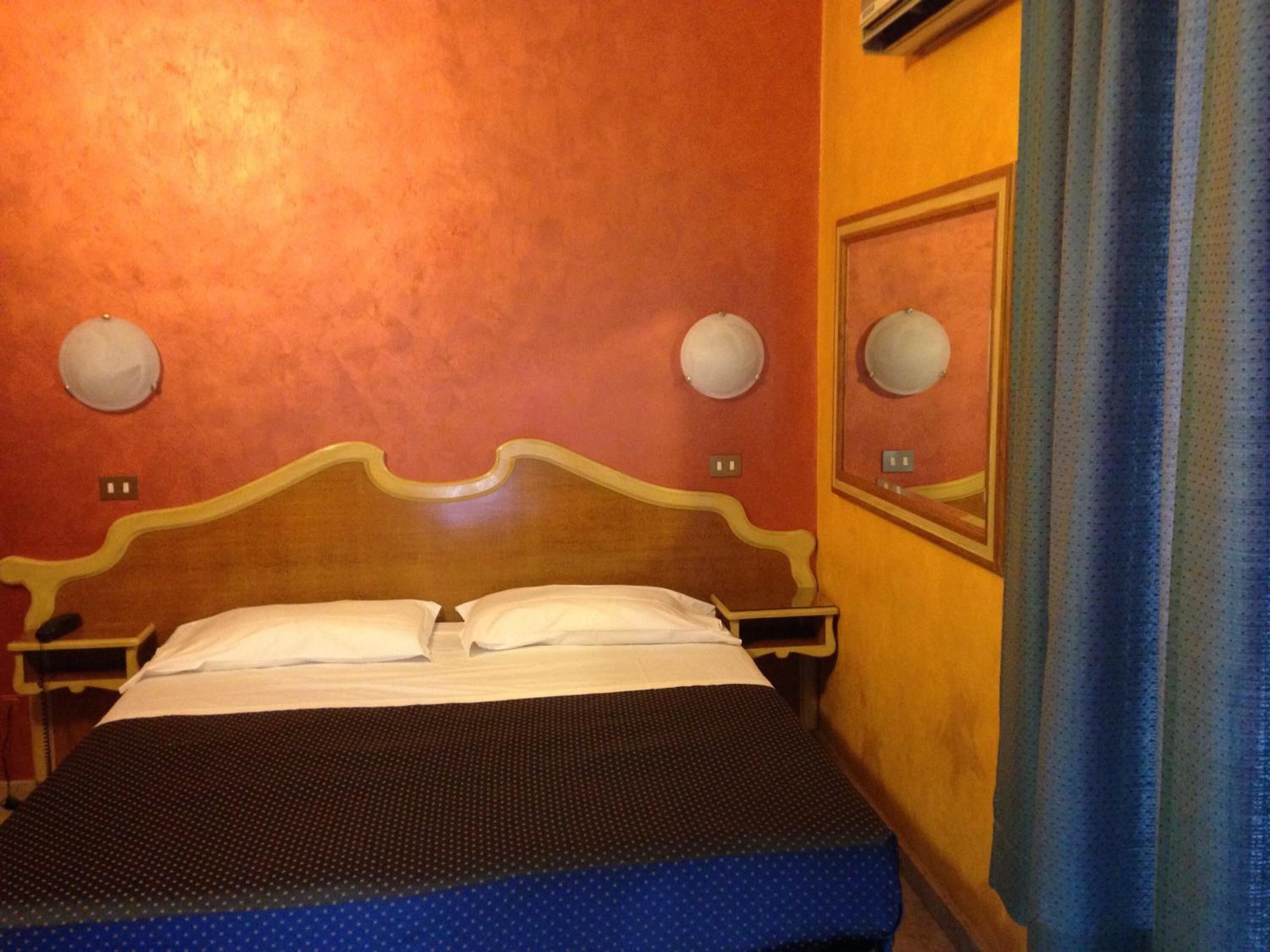 Una camera d'albergo con una testiera in legno, due comodini, un letto con lenzuola bianche e uno specchio a parete.