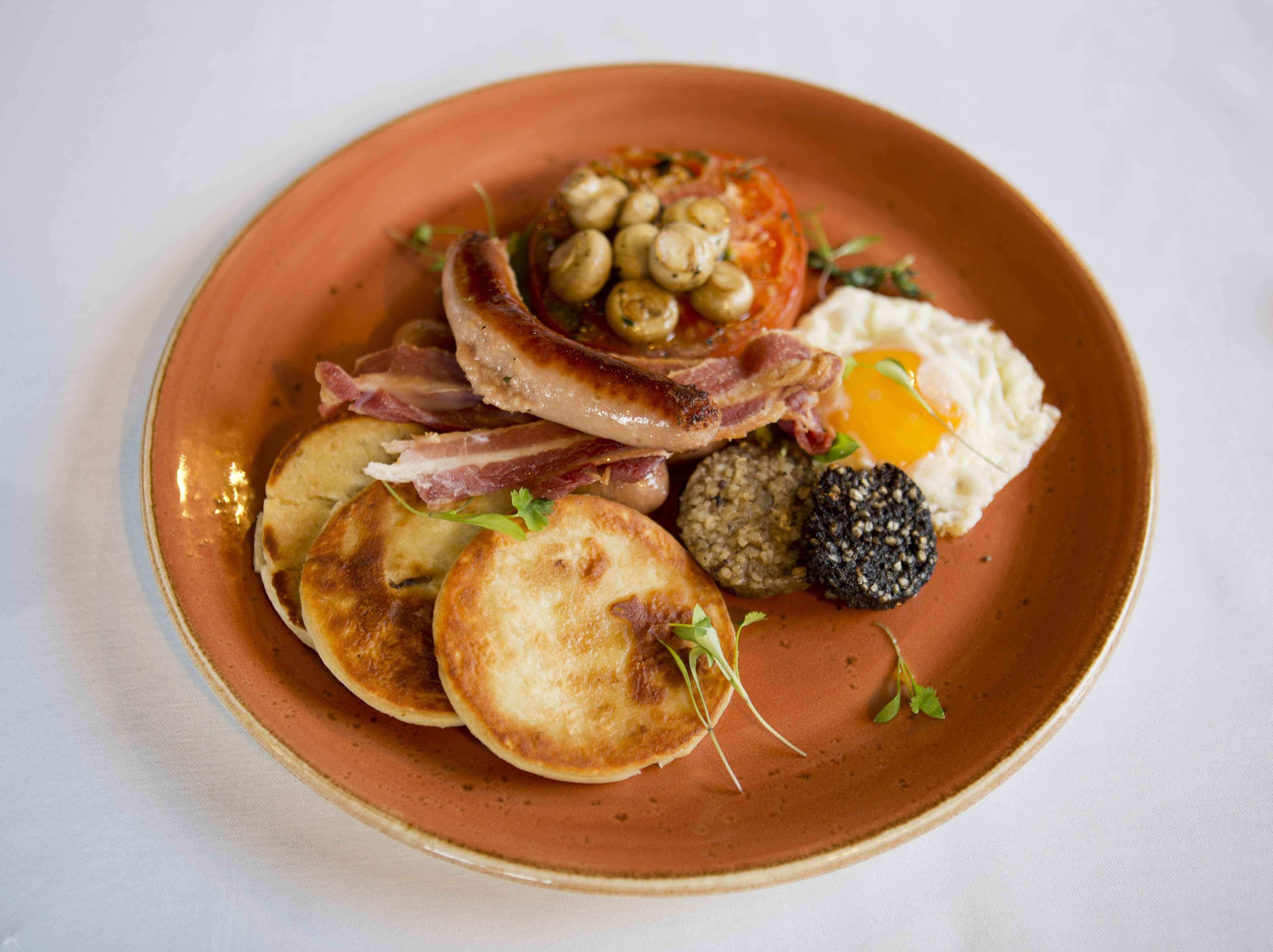 A plate of breakfast food including eggs, bacon, sausages, mushrooms, and black pudding.