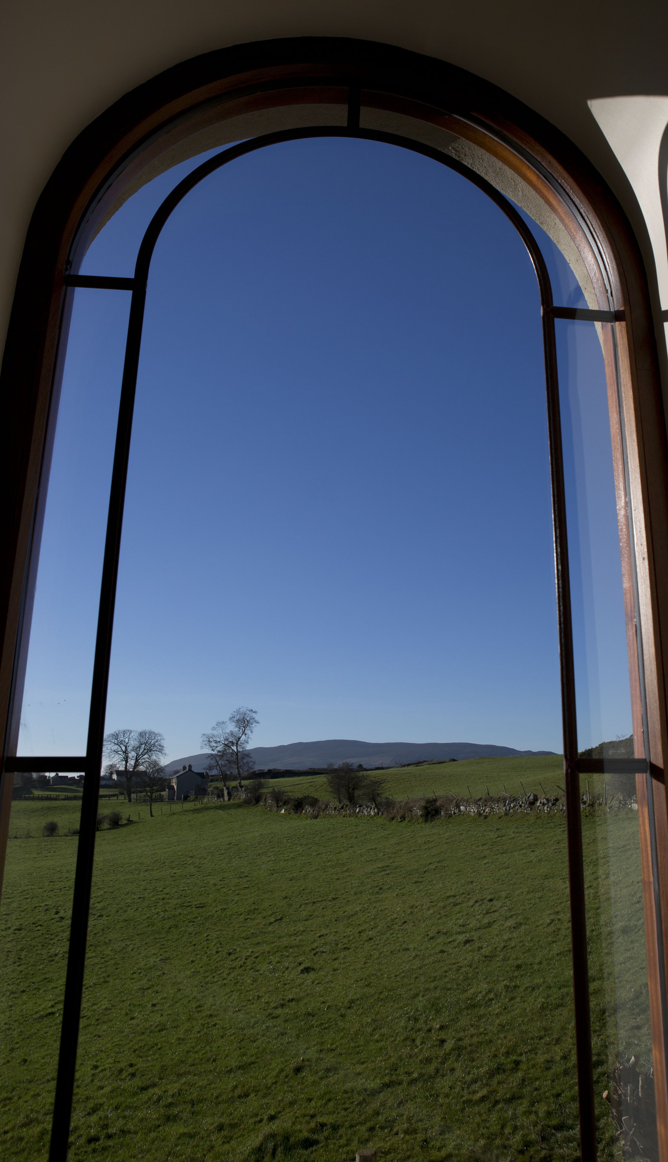A view through a large arched window of a green field, a stone wall, and distant hills.