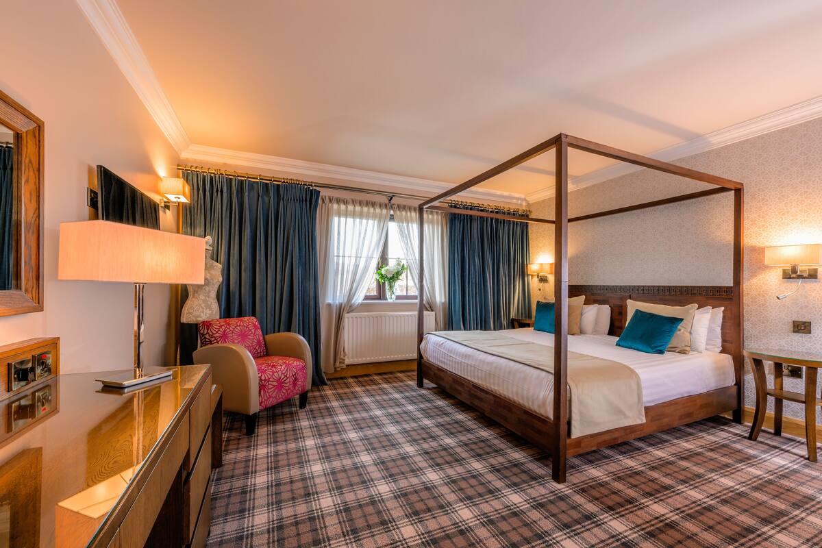 A four-poster bed with a plaid carpet, a desk with a lamp, and a chair with a floral pattern.