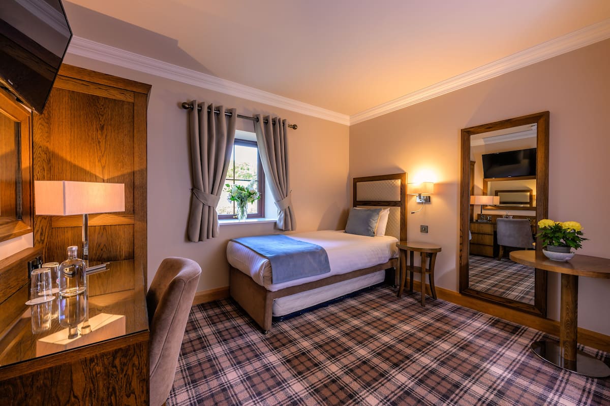 A hotel room with a bed, a desk with a lamp, a chair, a small table with a plant, and a mirror.