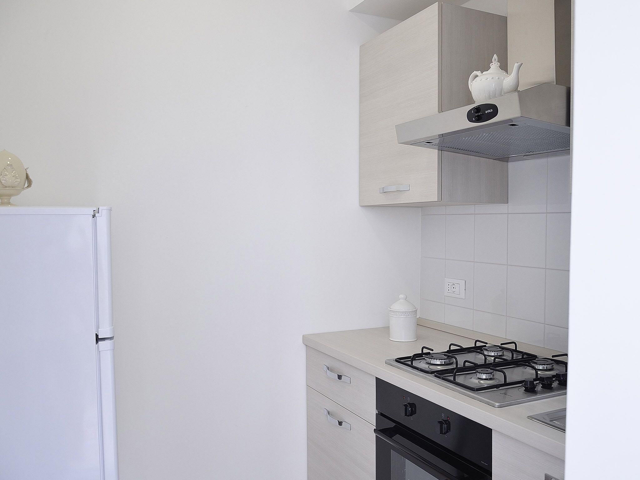 Una cocina con un refrigerador blanco, una estufa a gas y un microondas. Hay un armario blanco sobre la estufa y una pared blanca con un enchufe.