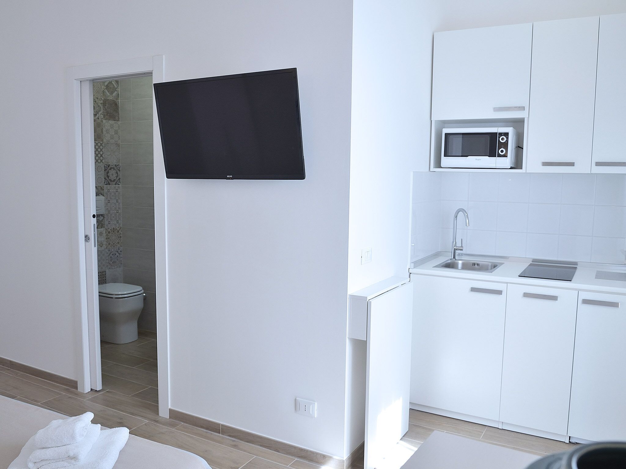 A compact kitchen with white cabinets, a microwave, and a sink. A black towel hangs on the door, and a toilet is visible in the adjacent bathroom.