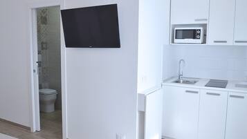 A compact kitchen with white cabinets, a microwave, and a sink. A black towel hangs on the door, and a toilet is visible in the adjacent bathroom.