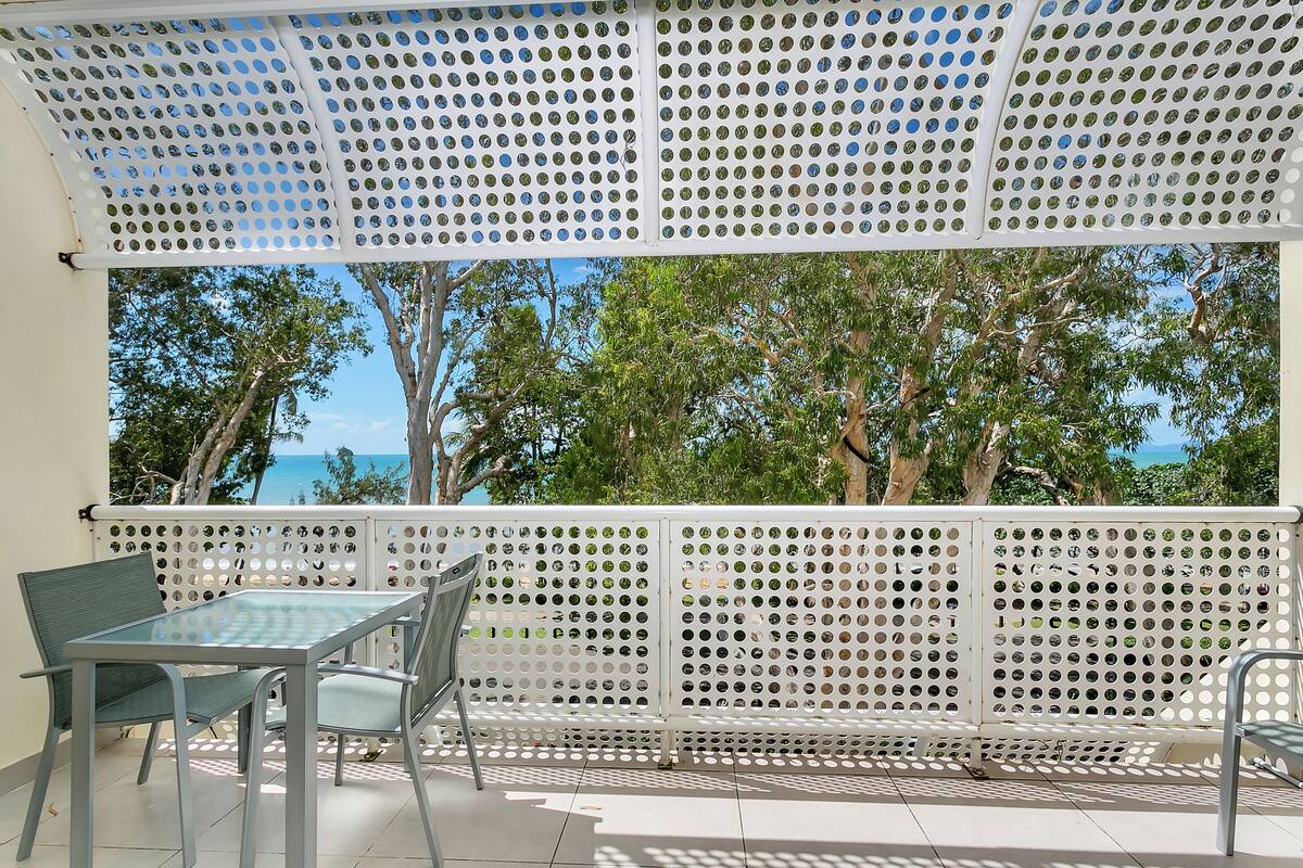 Un balcon avec une table et des chaises en verre, un paravent blanc ajouré et une vue sur les arbres et la mer.