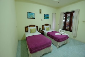 A hotel room with two beds, each with a purple bedspread and white pillows. There are two framed pictures on the wall, a window with curtains, and a small table between the beds. - Aonang Friendly Bungalow (Krabi)