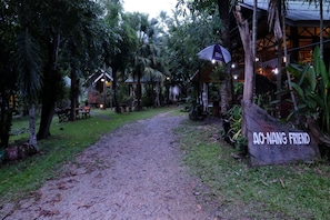 A rustic pathway leading to a wooden structure labeled "AO-NANG FRIEND" surrounded by lush greenery and a thatched-roof building. - Aonang Friendly Bungalow (Krabi)