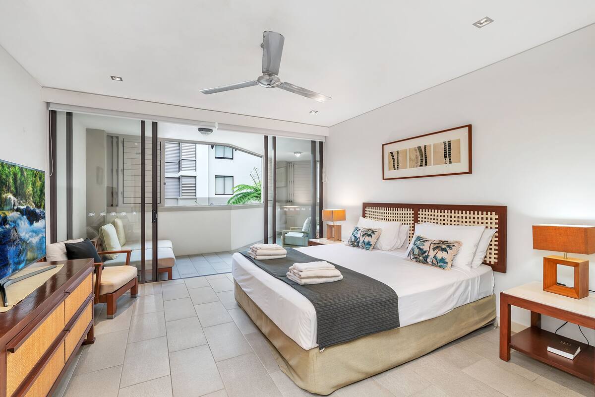 A modern hotel room with a large bed, a ceiling fan, a sliding glass door leading to a balcony, and a painting on the wall.