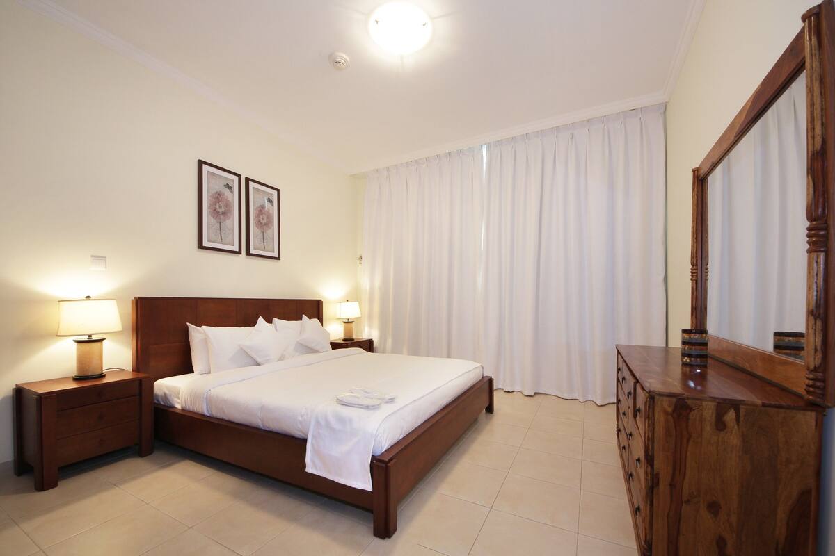 Une chambre d’hôtel bien aménagée, avec un lit en bois, deux lampes de chevet, une commode en bois et un grand miroir.