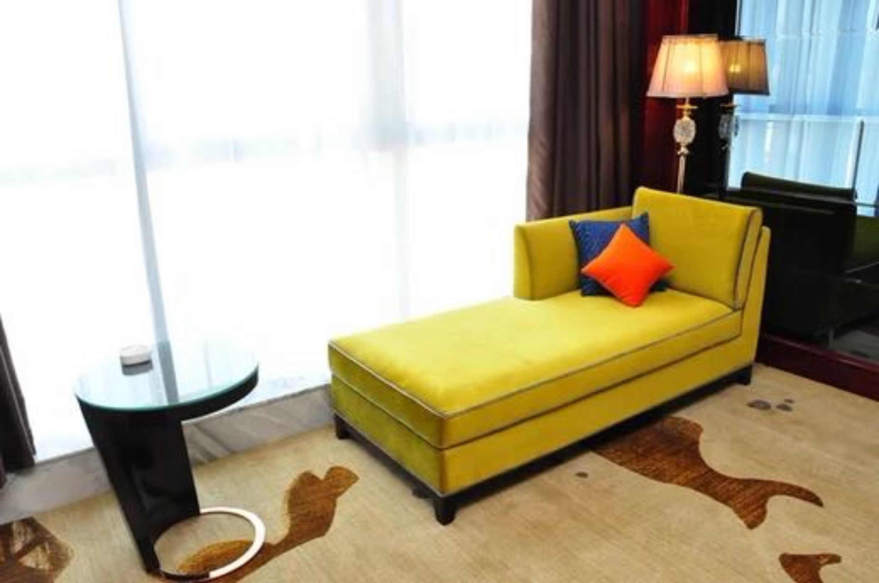 Un sofá amarillo con un cojín azul y naranja en una habitación de hotel, con una mesita de vidrio y una lámpara.