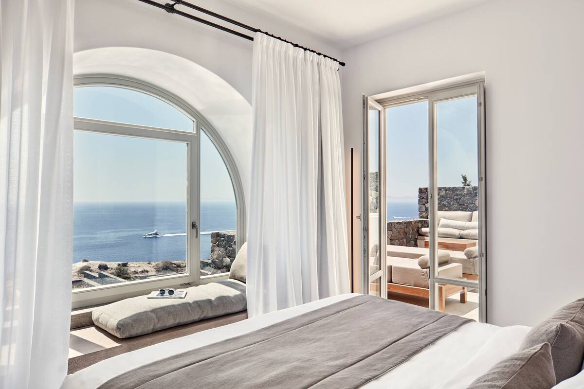 A bedroom with a large window overlooking the sea, a balcony with seating, and a bed with a view.