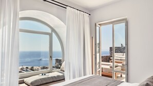 A bedroom with a large window overlooking the sea, a balcony with seating, and a bed with a view.