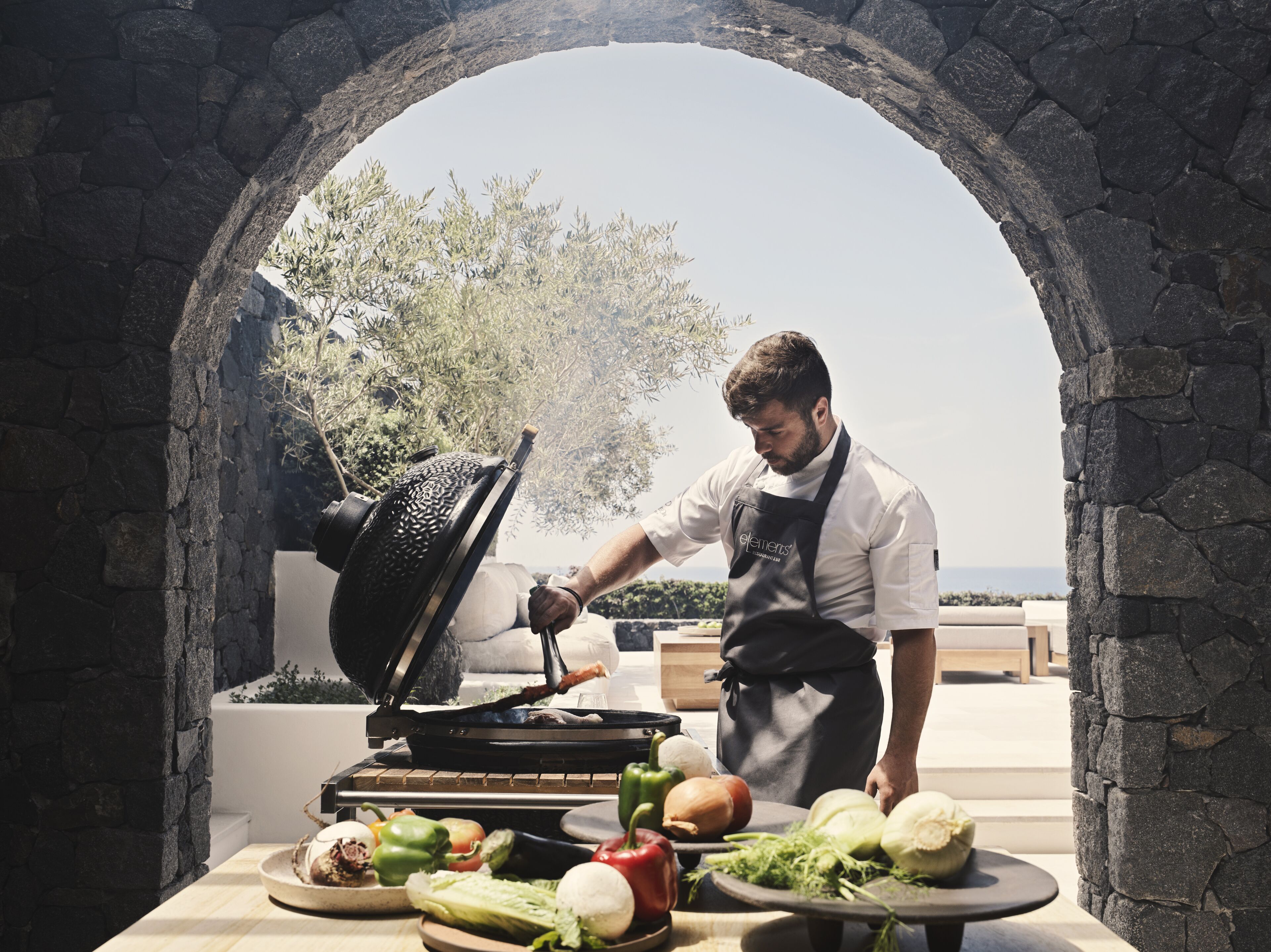Un chef cuisinant des grillades à l’extérieur, sous une arche en pierre, entouré de légumes frais.