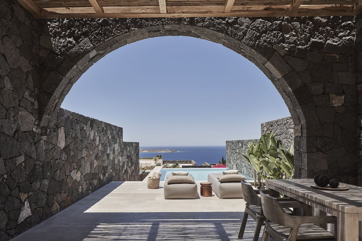 Un arc en pierre mène à un espace piscine agrémenté de chaises longues et d’une table, offrant une vue sur la mer.