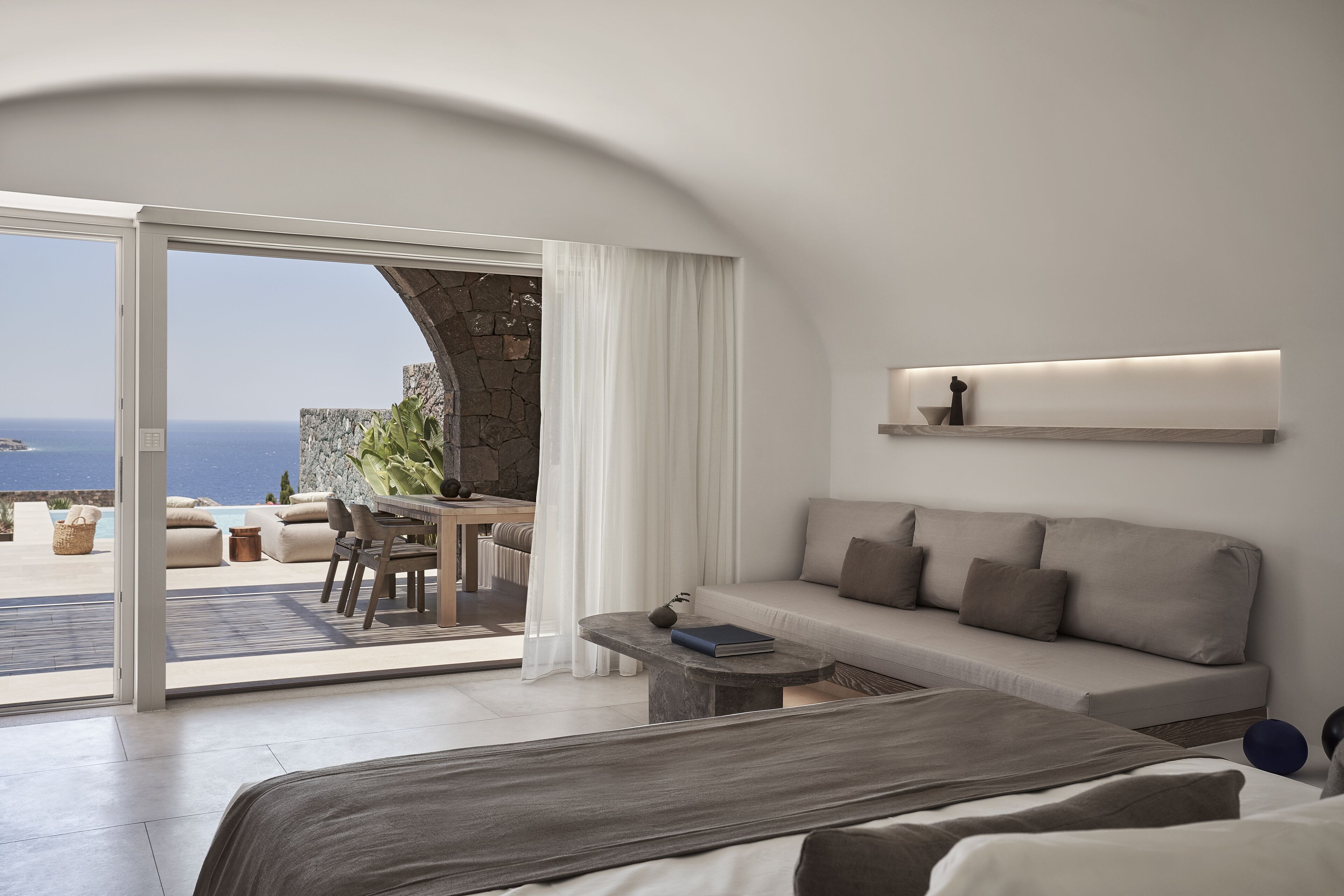 A modern bedroom with a large bed, a sofa, and a dining area with a view of the sea.