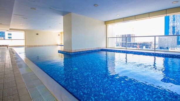 A modern indoor swimming pool with clear blue water, a tiled edge, and a view of a cityscape through large windows.