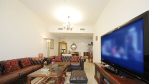 A spacious living room with a large flat-screen TV, leather sofas, a glass coffee table, and a chandelier.