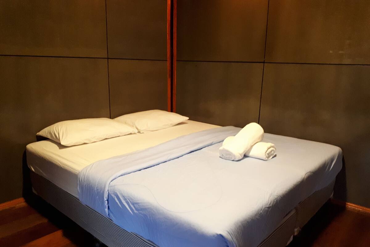 A neatly made bed with white and blue bedding, a wooden headboard, and a pair of rolled towels on the foot of the bed.