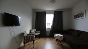 Een woonkamer met een flatscreen televisie aan de muur, een gestreept fauteuil, een donkere bank, een kleine ronde tafel met een bloemen tafelkleed en een raam met gordijnen.