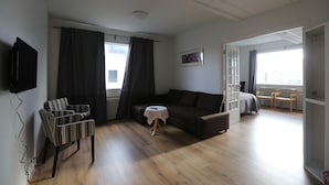 Een moderne woonkamer met een bank, een kleine ronde tafel, een aan de muur bevestigde televisie en een raam met gordijnen.