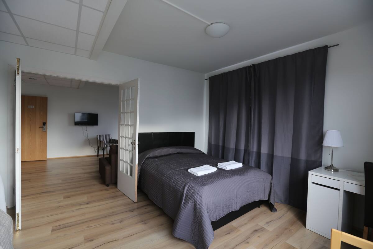 Moderni hotellihuone, jossa on puulattia, tumma päätyosaan varustettu sänky, työpöytä lampun kanssa ja seinälle asennettu televisio.
