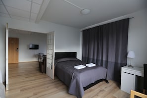 A modern hotel room with a wooden floor, a bed with a dark headboard, a desk with a lamp, and a television on the wall. - 100 Iceland Hotel (Reykjavik)