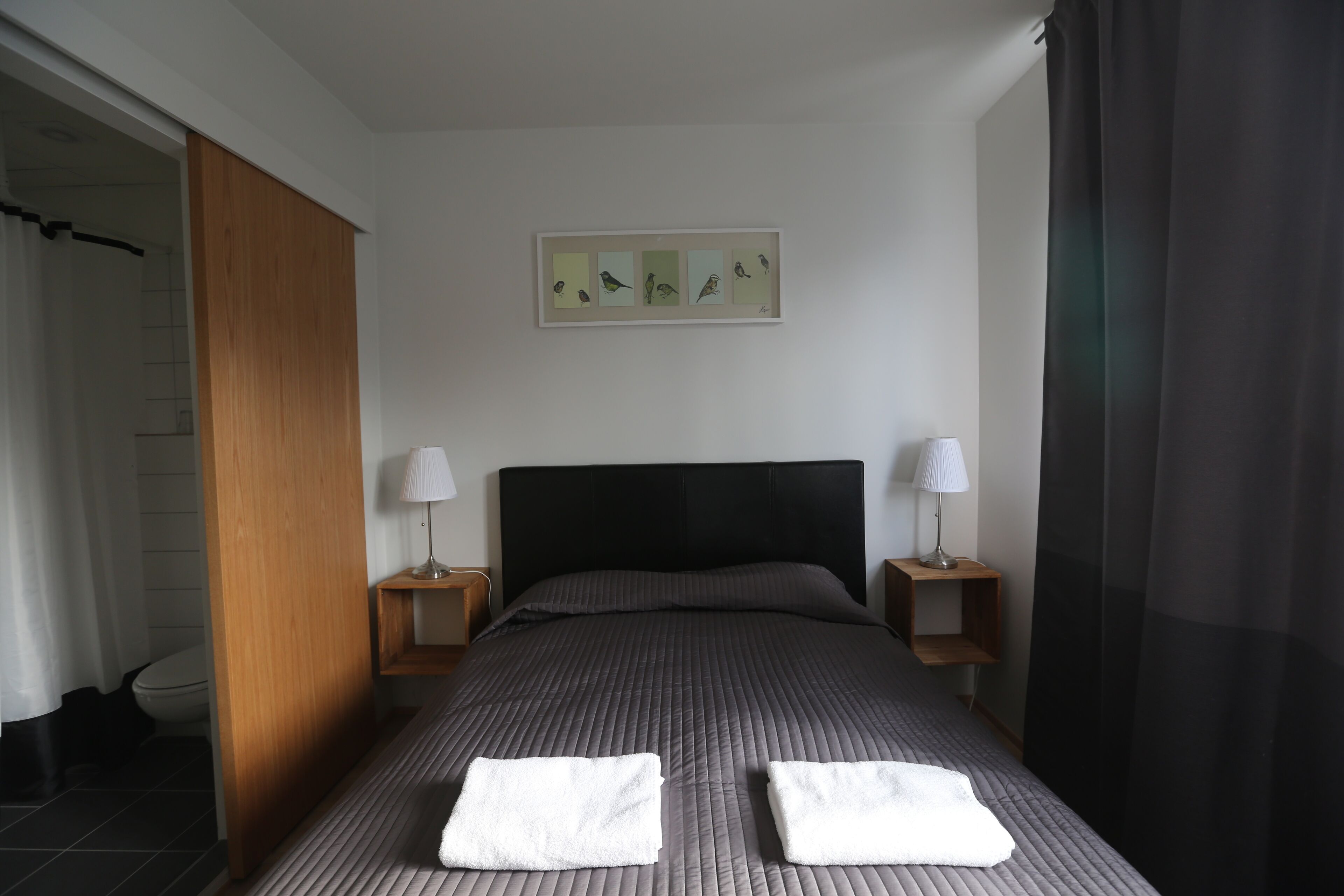Una habitación de hotel con una cama, dos mesitas de noche con lámparas, una puerta de madera, un cuadro enmarcado en la pared y un baño visible a través de una puerta abierta.