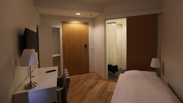 Een hotelkamer met een houten vloer, een bed met een witte dekbedovertrek, een bureau met een lamp, een stoel, een televisie en een deur naar een badkamer met douchegordijn.