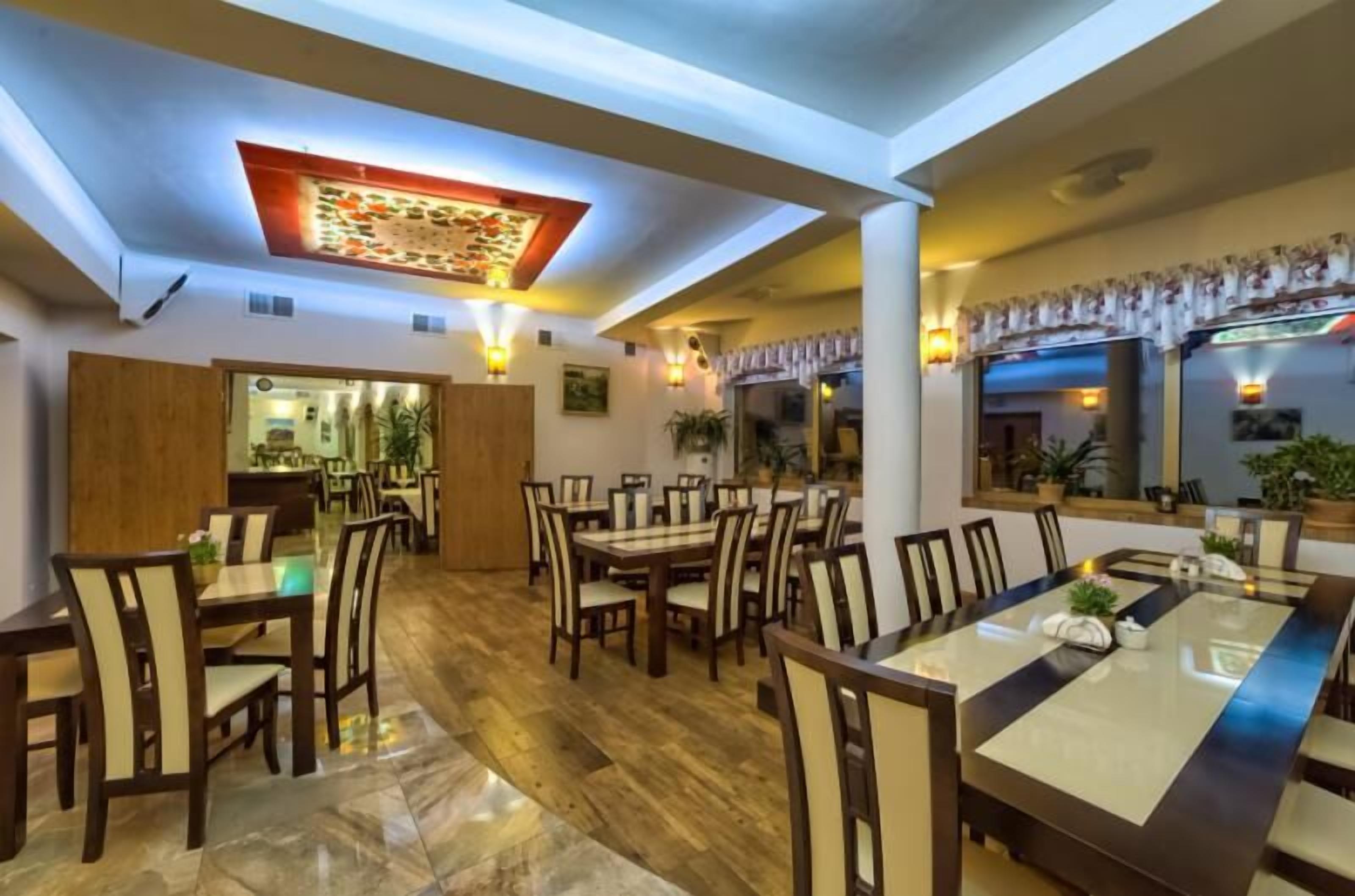 A spacious dining area with multiple tables and chairs, a wooden floor, and a ceiling with a decorative light fixture.