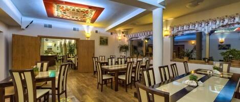 A spacious dining area with multiple tables and chairs, a wooden floor, and a ceiling with a decorative light fixture.