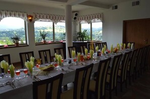 A dining area with a long table set for a meal, large windows with a view of the countryside, and a wooden door in the background. - Montenero Resort & SPA (Bukowina Tatrzanska)