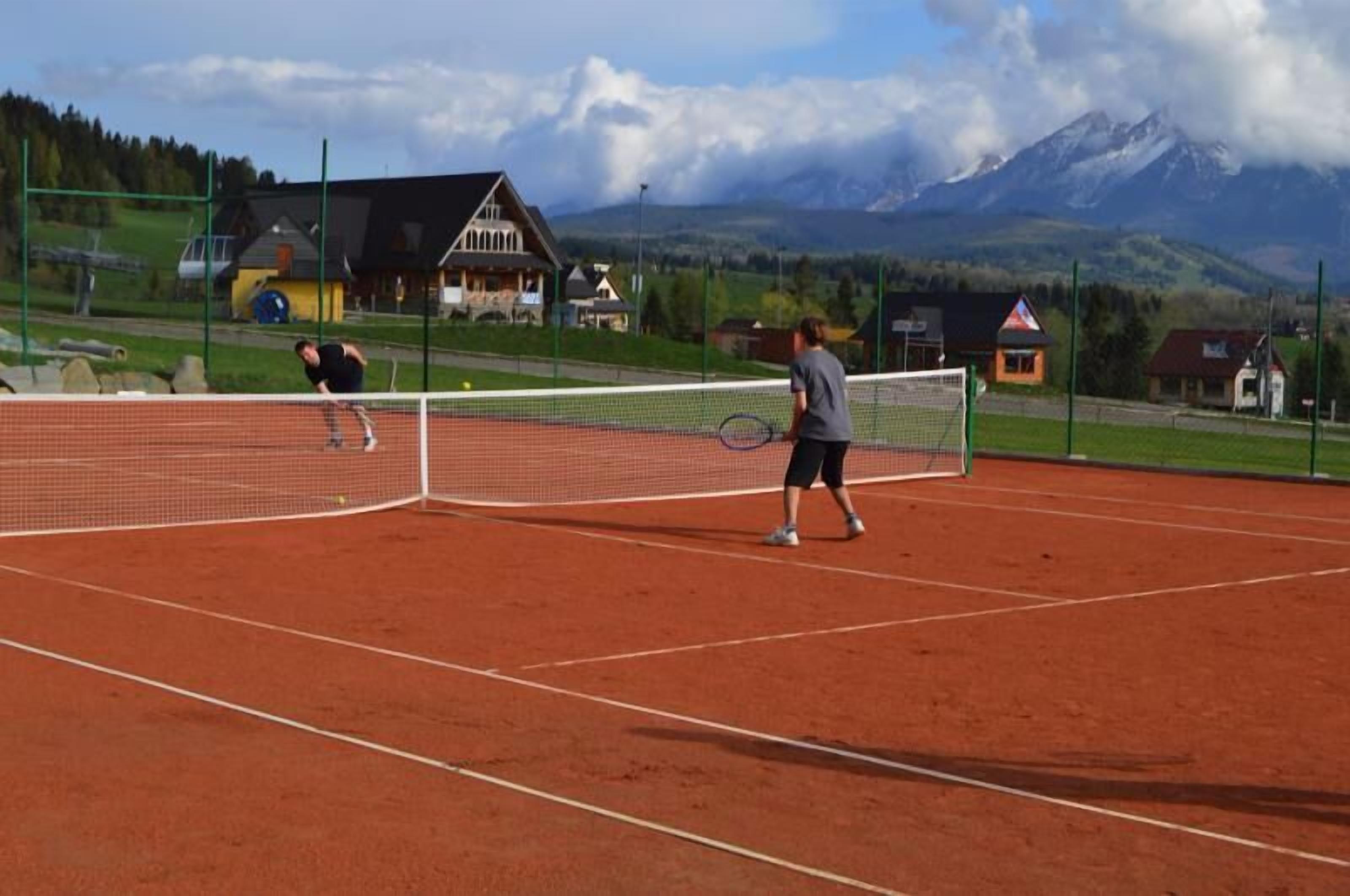 Una cancha de tenis con dos jugadores, rodeada de casas y montañas al fondo.