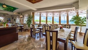 Un comedor amplio con varias mesas y sillas, ventanas grandes con cortinas y un mostrador de recepción.