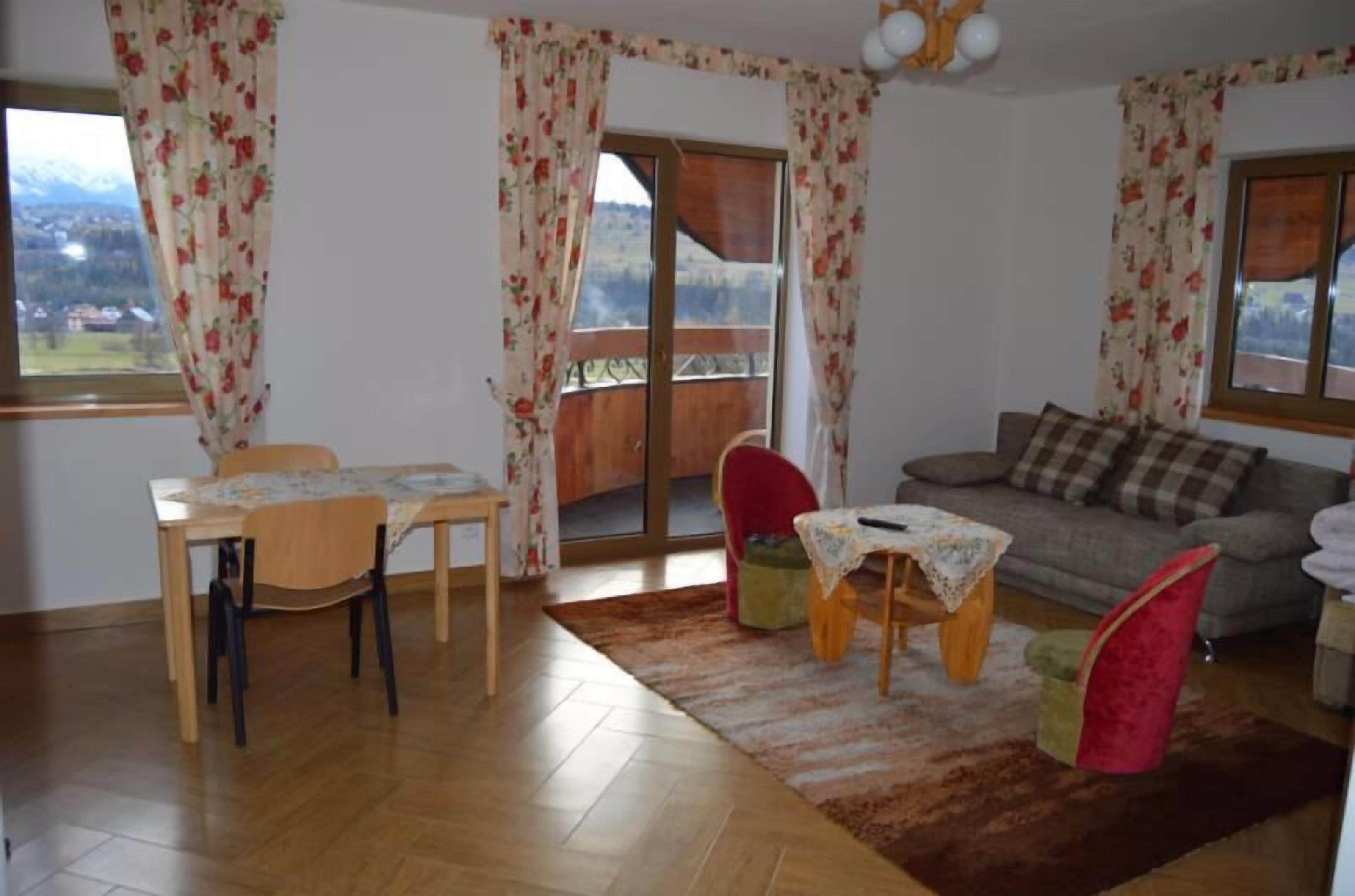 Ein Wohnzimmer mit Holztisch, zwei Stühlen, einer Couch und einem kleinen Hocker. Vor den Fenstern hängen Vorhänge und es gibt einen Blick auf einen Balkon mit Holzgeländer.